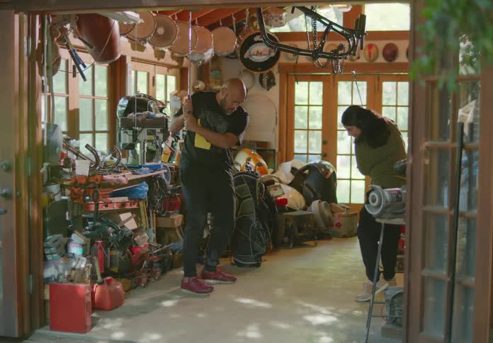 A father and daughter stand across from each other in a windowed storage room full of equipment as he teaches her golf swing technique.