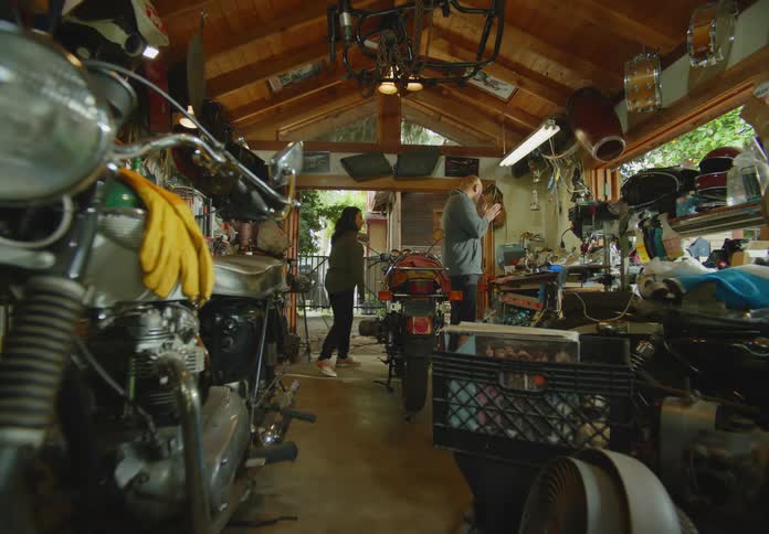 Unique POV from the front end of a motorcycle into the workshop beyond where a man and woman are working on another motorcycle and give each other a high-five once completed and then move on to another project.