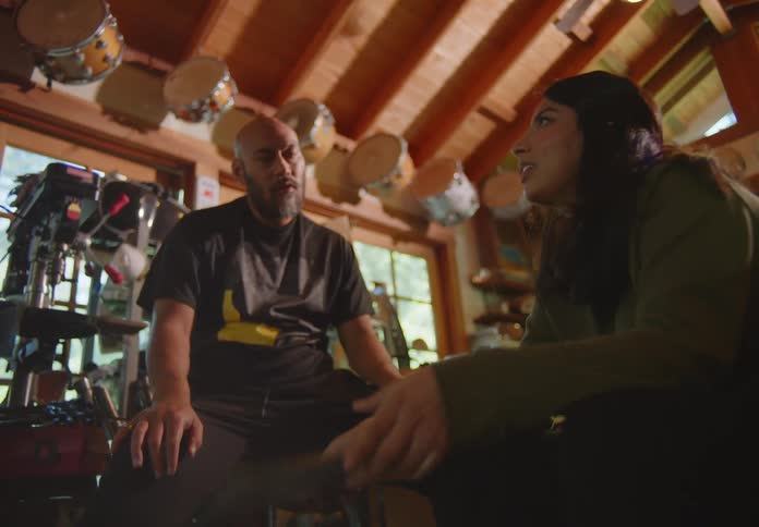 An ethnic couple sit in a cluttered storage room filled with various items and the woman holds a vinyl and laughingly makes a snarky remark as the man snatches the record from her while jokingly feigning outrage.