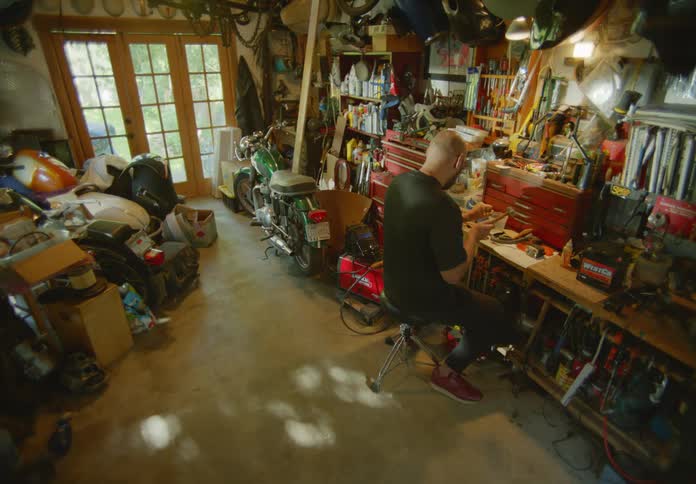 Birds eye view of a bald man with his back to the camera sitting on a stool at his workbench in a cluttered workshop with a hammer and chisel carving something.