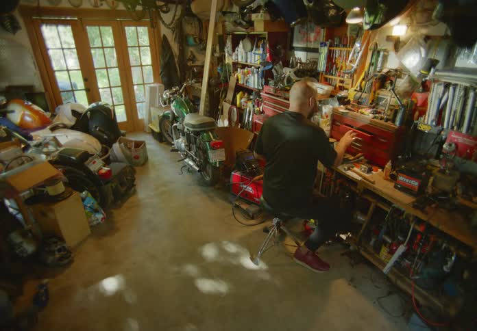 A bearded and bald man in a black t-shirt is working on a project in a cluttered garage workshop filled with various tools, equipment and hobby items.