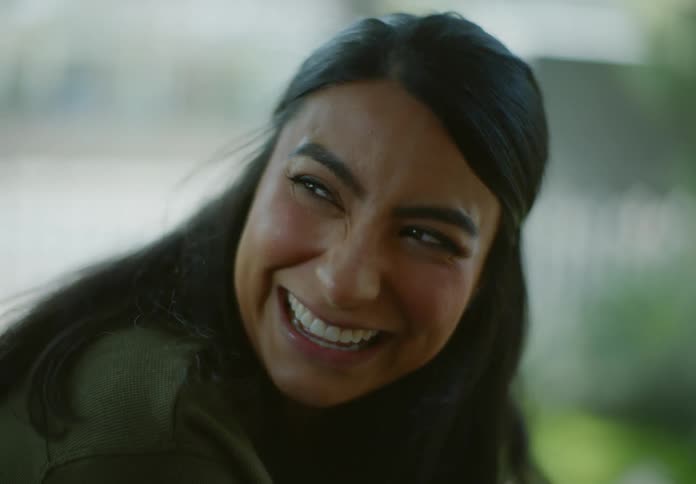 A woman with black hair and a green sweater laughs out loud while drinking a glass of water in the yard