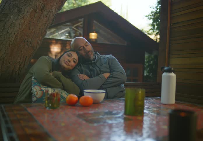 A woman with dark hair and a green shirt resting her head on a bearded mans shoulder while they both sit at a table in the yard