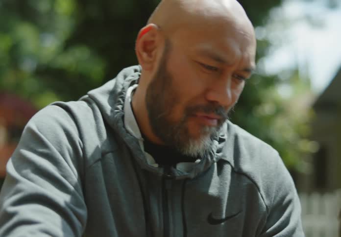 A bald, tanned, bearded man in a hoodie stands outside with a dark-haired woman and they smile and chat as they examine something they are looking at below