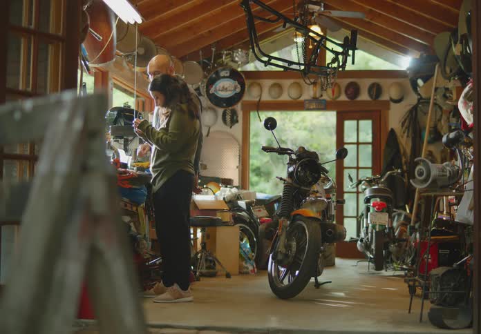A man and woman look at and examine items on the workbench of a cluttered garage with natural light from windows