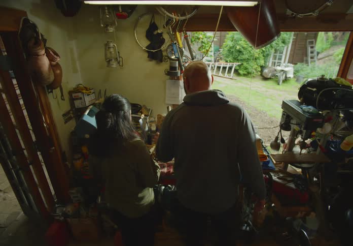 A bearded and bald man with a hoodie and a dark-haired woman enter a cluttered workshop garage together and start examing and gathering parts from a table full of items.
