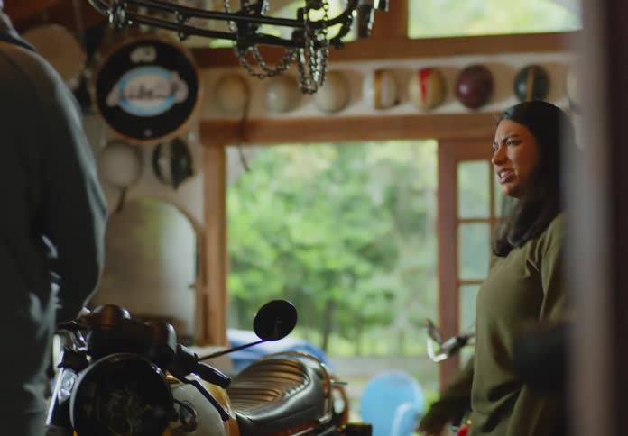 A bald, bearded man and a young dark-haired woman walk into a cluttered workshop and look around as the man rests his hand on a motorcycle and looks down at it.