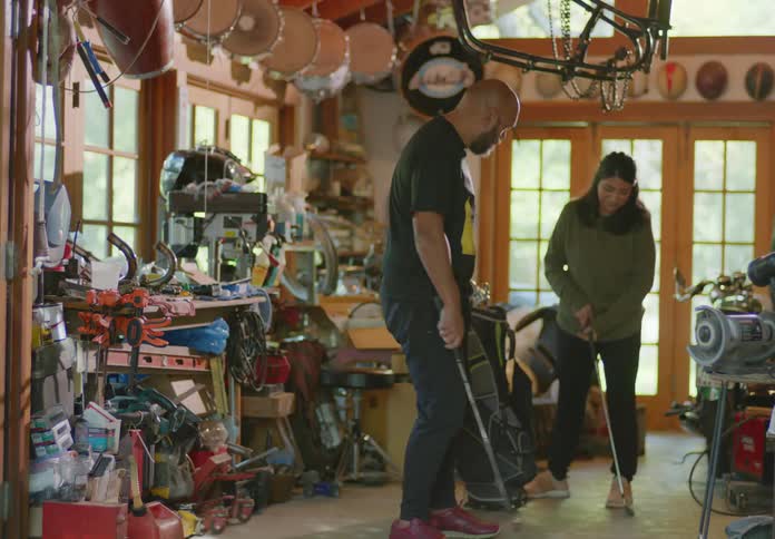 A father and daughter stand in a cluttered garage holding golf clubs as he teaches her technique on how to swing.
