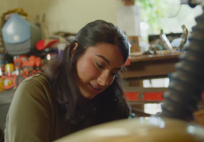 A young, ethnic woman talks to someone offscreen as she polishes an object in a workshop full of tools.