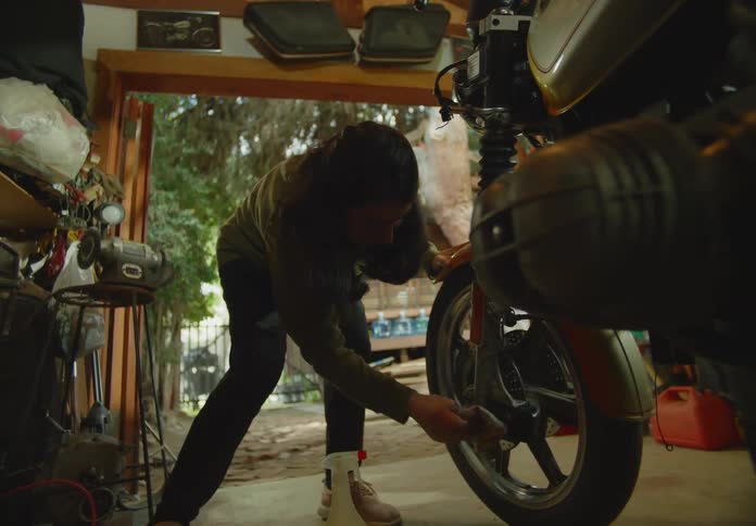 A young, ethnic woman in an outdoor workshop is bent over wiping down the tire of a motorbike with a rag.