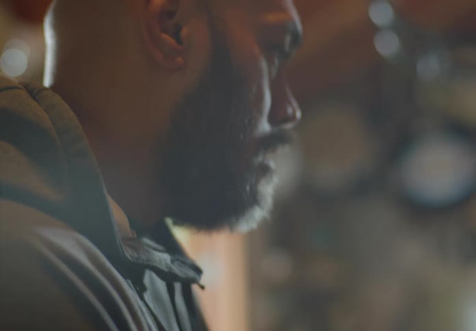A olive-skinned man with bald head and beard looks down and focuses on a task intently as he mumbles something to a person offscreen.