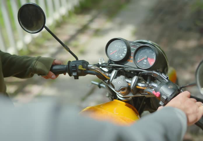 View of a motorcycle instrument panel from over the shoulder of a man pointing to the ignition switch and explaining how to turn the bike on to a woman standing beside him.