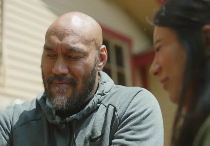 A tan and bald man with a beard explains something to a dark-haired woman beside him who smiles and squints her eyes, then raises her eyebrow as she opens them, looks down, smiles and giggles.