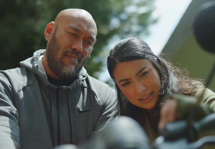 A bald, Polynesian man with facial hair sits on his motorcycle next to a young, ethnic woman who leans in and twists a dial with a puzzled and focused look as they discuss the result and she smiles.