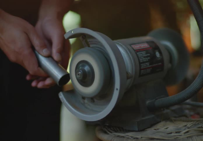 A man shaves a metal pipe with a spinning grinder machine creating friction sparks as he slowly turns the pipe.