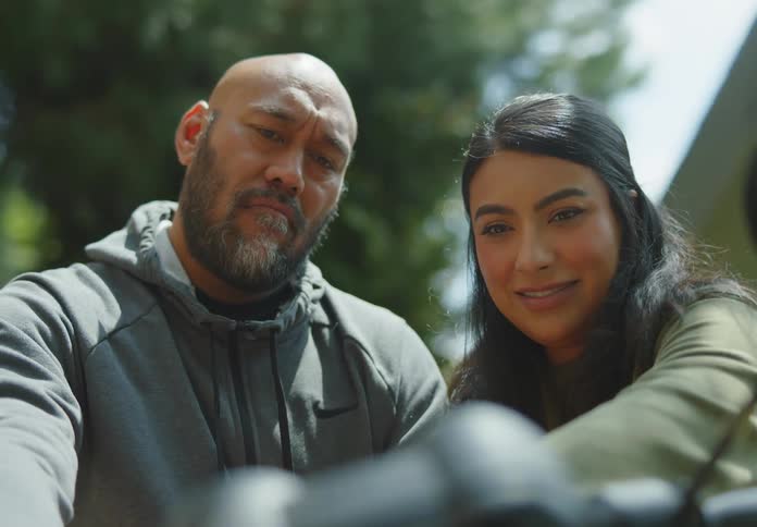 A tan and fit, bald male Pacific Islander grips the handlebar of his motorbike and explains the mechanics of it to a young, dark-haired female who is leaning in over his shoulder and touching something on the instrument panel.
