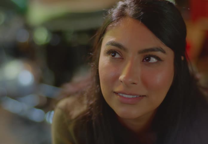 A seated young, ethnic woman with almond-shaped dark eyes looks up at someone who has just asked her a question, to which she squints her eyes, replies, smiles and tucks her chin in a laugh.