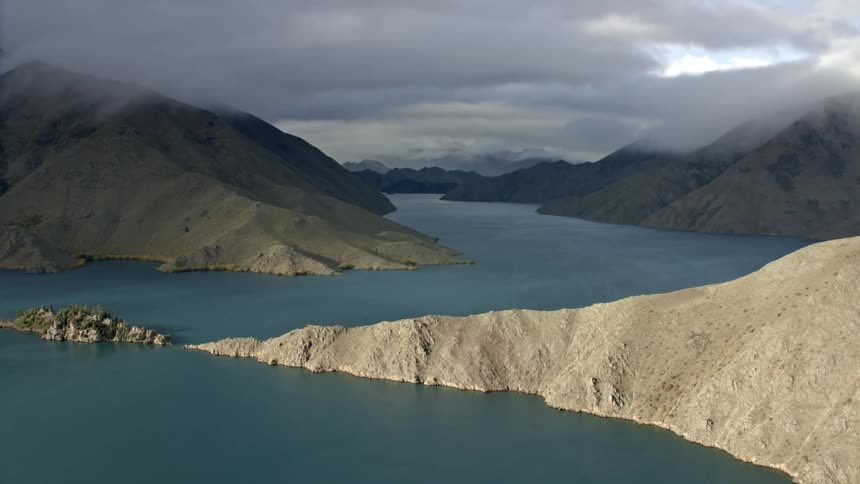 Sunny foreground hills with a large lake surrounded by rugged mountains under a dramatic moody sky in the distance