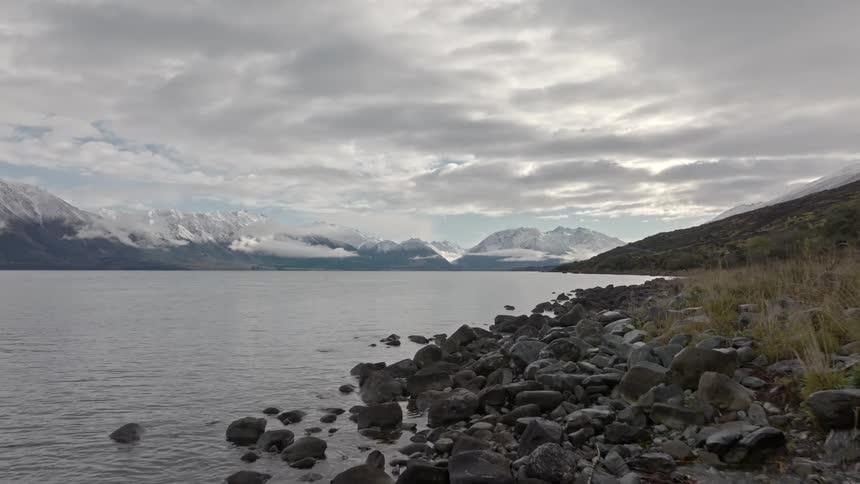 Very low level aerial across a small stream of water that goes into the lake and in the background there is a mountain range