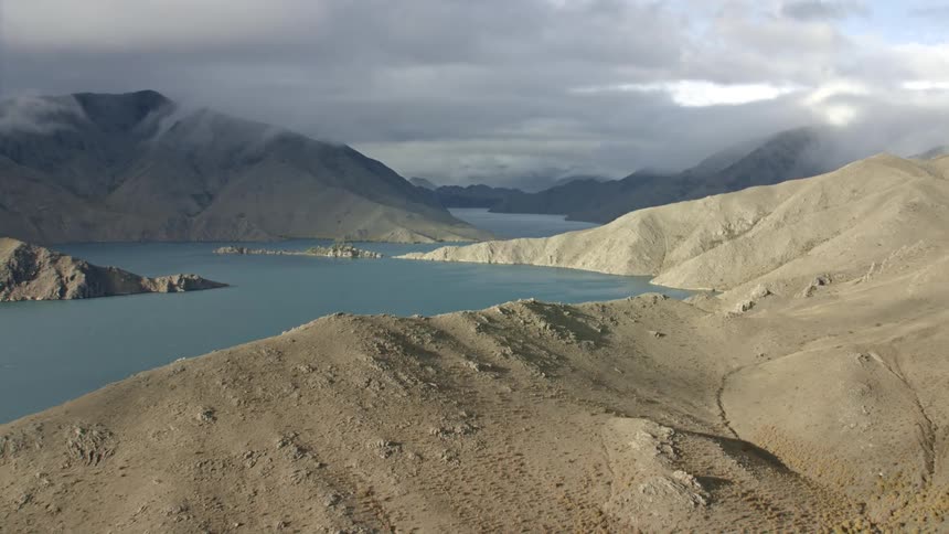 Aerial view of a serene lake with a small island surrounded by hills and mountains under a cloudy and moody sky