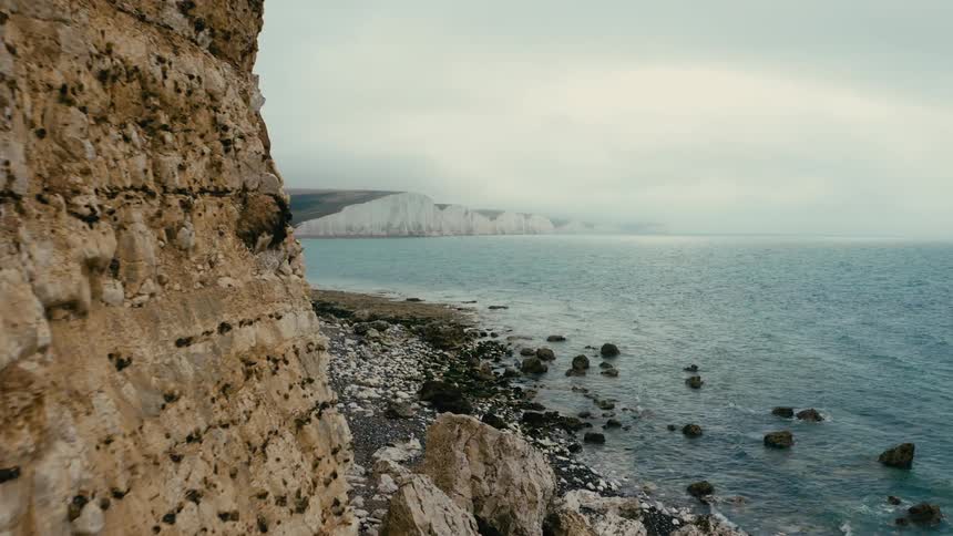 A coastal cliff a pebble beach and white cliffs in the distance under an overcast sky