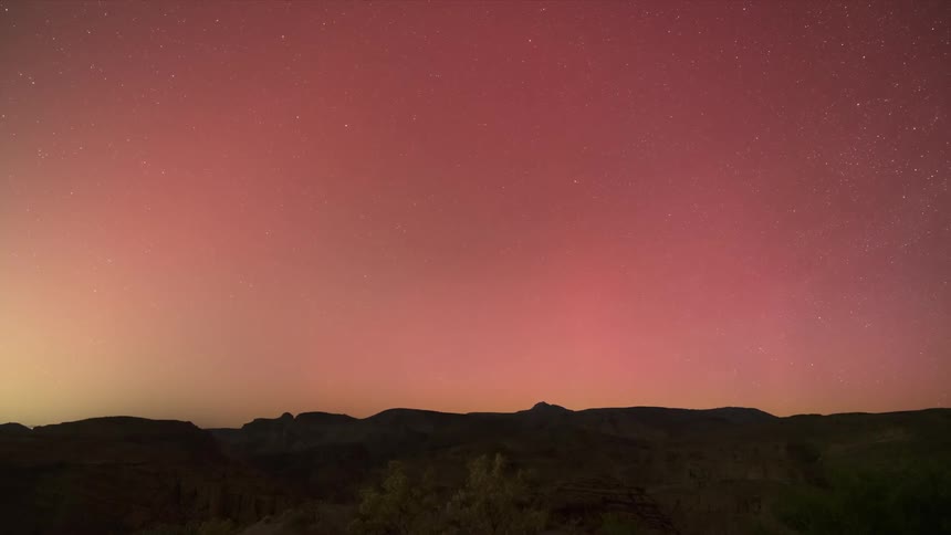 A gradient of pink and purple aurora in the night sky above a dark silhouette of mountainous terrain