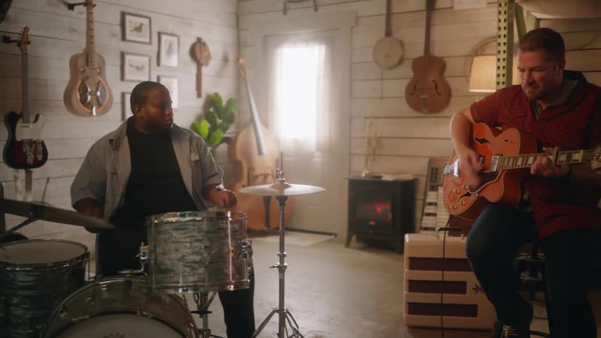 Two musicians in a room with one playing the drums and the other a guitar