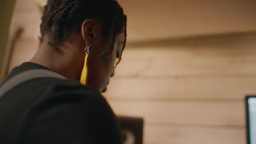 A woman with dreadlocks and yellow earrings clad in a black shirt nods intently at the computer screen in a recording studio
