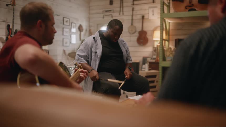 Three people playing music in a room with one man playing guitar and another using drum sticks on his leg