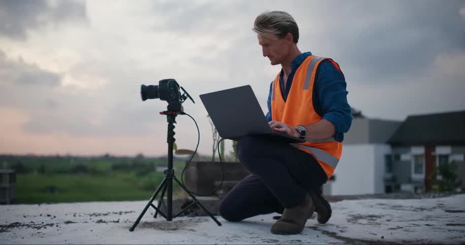 A man in an orange vest kneels atop a building roof with a camera and laptop