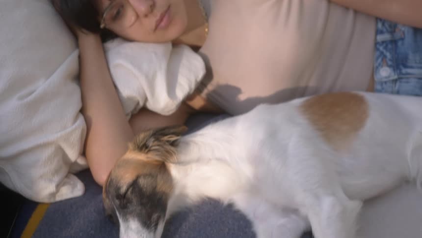 A woman changes channels with a remote while her dog sleeps next to her