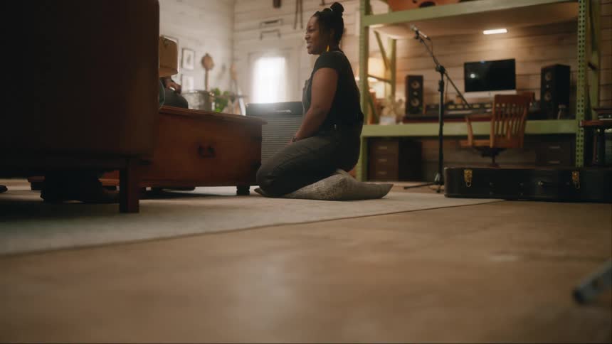 Woman sitting on a cushion singing and next to her a person playing the drums while they are in a studio
