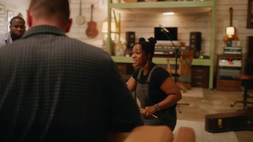 Four individuals in a room filled with musical instruments with one holding a guitar and a woman singing