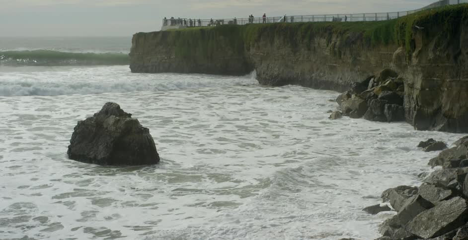 Ocean waves crashing into rocks while people observe on cliff above