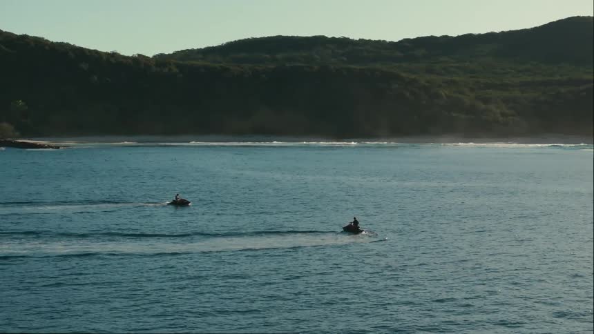 Two jet skis race across a turquoise ocean kicking up spray
