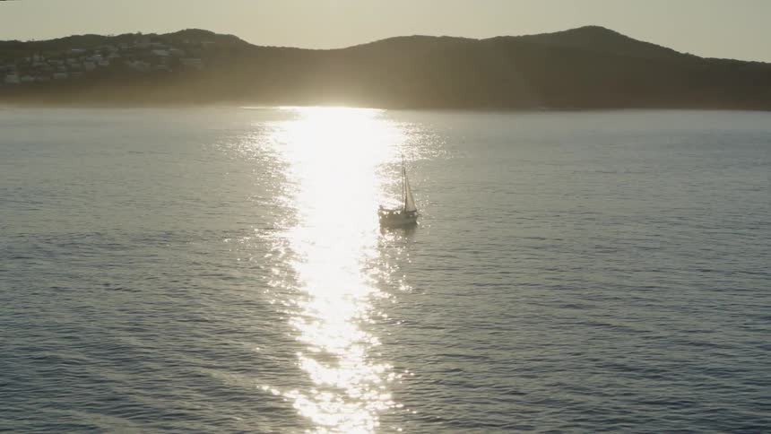 A lone sailboat glides atop the surface of the ocean with the shoreline in sight