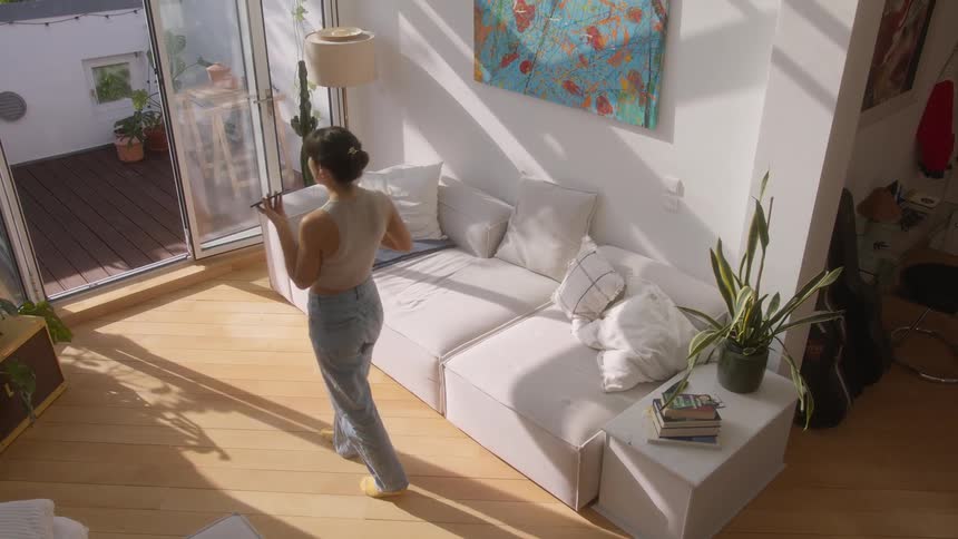 A woman wearing a beige top and jeans talks on speakerphone on the phone in the living room at home