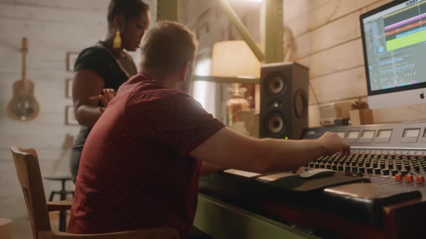 A bearded man in a red tshirt collaborates with a woman in a jumpsuit in a professional recording studio