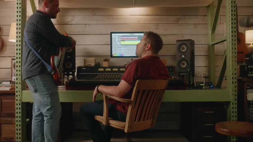 Man holding a guitar talking to a colleague next to him in front of a music mixer in a studio