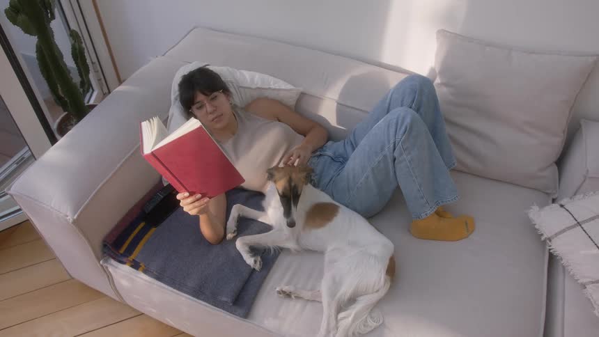 A woman lies on a couch reading a book with a dog on her lap