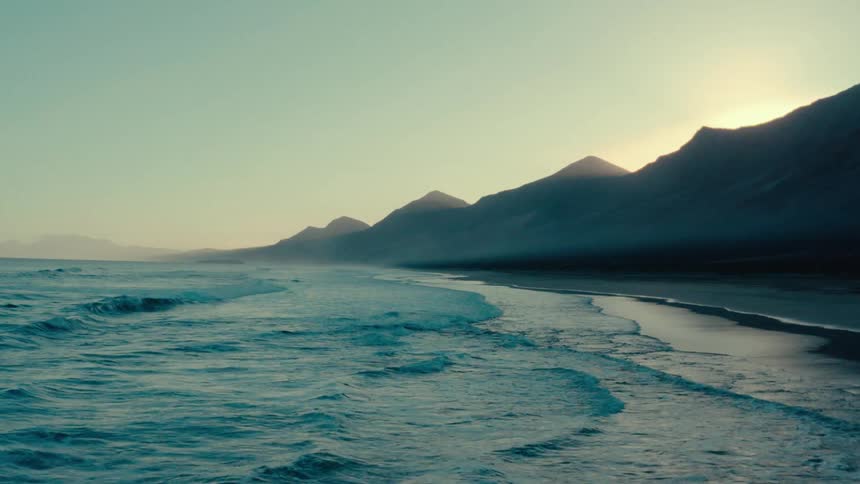 Ocean waves crashing against the shore with mountain range in the background at sunset
