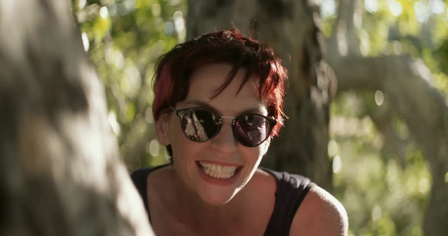 Smiling woman with sunglasses peeking around from a tree and waving