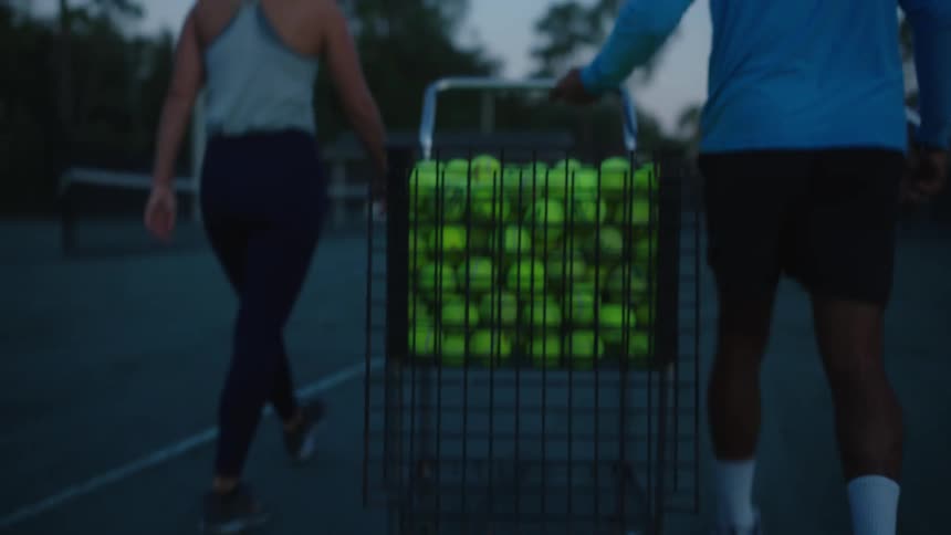 A man walks with a woman on a tennis court while pulling a cart full of tennis balls