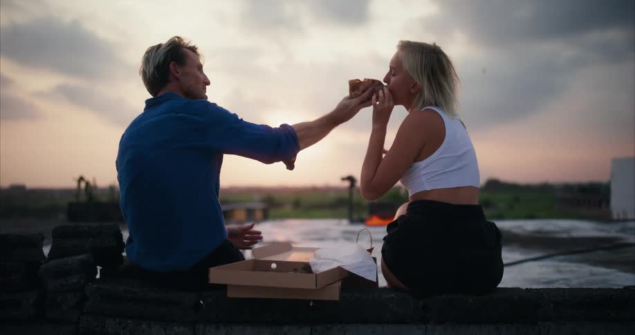 Man feeding a piece of pizza into the mouth of a woman next to him while they are outdoors in the late afternoon