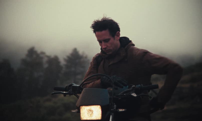 A man sitting on a motorcycle in the countryside takes out his helmet