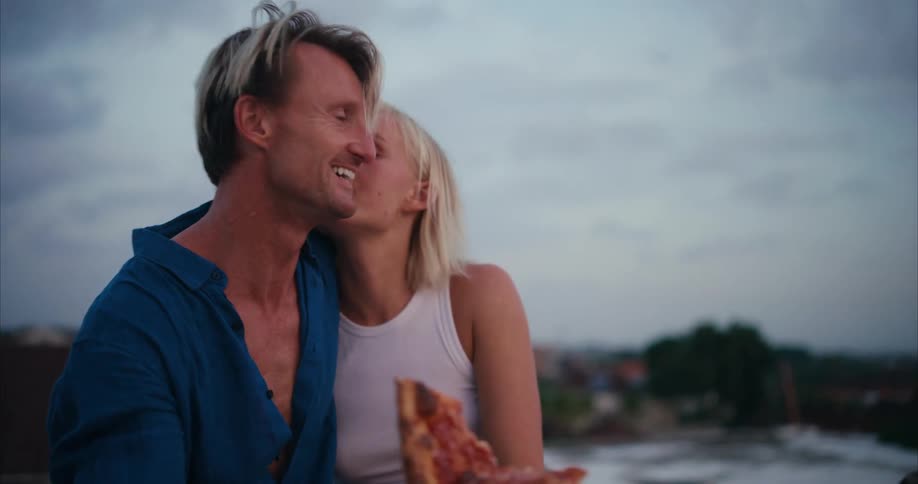 Man and woman hugging side by side in an openair environment eating pizza
