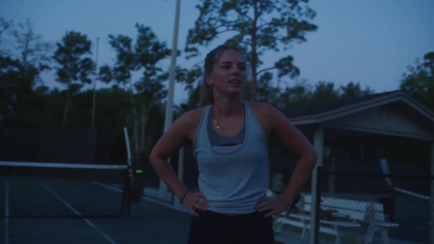 A tired woman in a tennis court stretching her arms behind her head after doing exercise