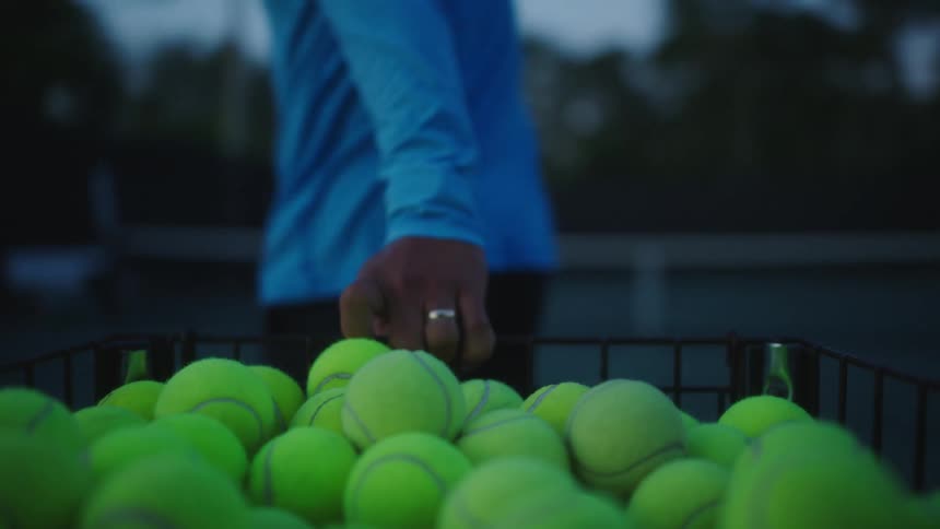 A person pulling a basket of tennis balls on a tennis court during dusk