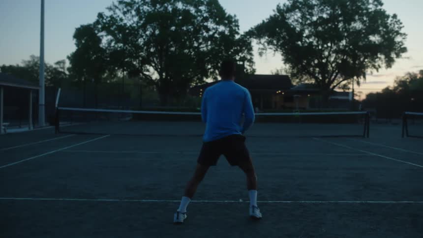 Two people on a court in the late afternoon playing tennis