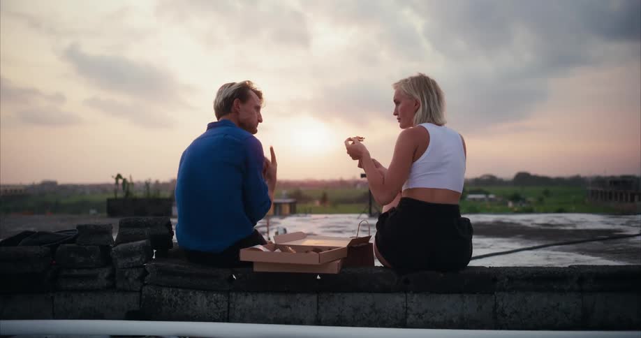 A man and a woman sitting on a rooftop eating and feeding each other pizza